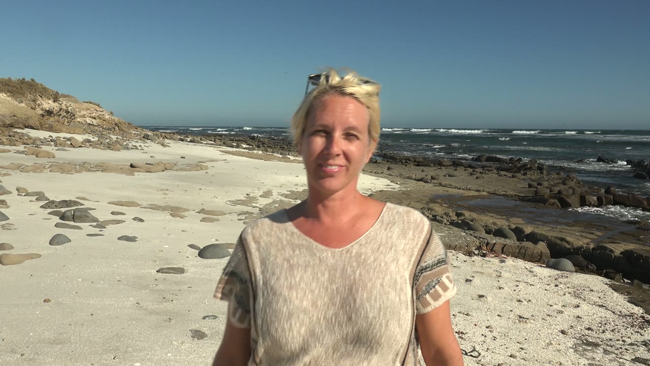 A blonde woman faces the camera on a sandy La Bocana beach with scattered stones, dark shoreline rocks, blue ocean waves, and a clear sky behind her.
