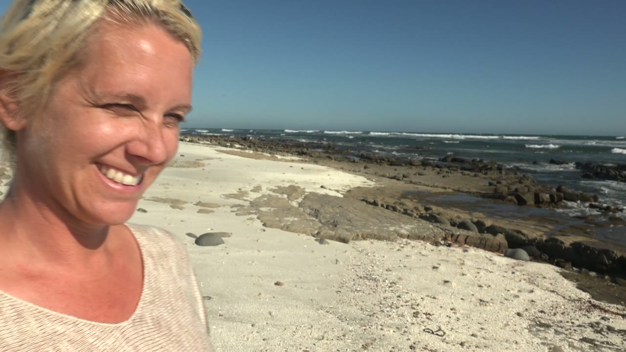 A close side view catches the smiling woman moving out of frame at La Bocana, with the sandy beach, dark rock line, surf, and clear horizon still visible.