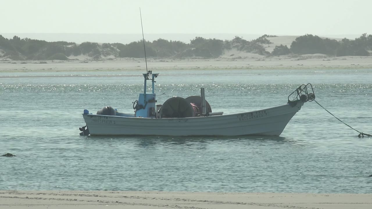 At 0:00, a small white fishing boat rests in shallow blue-green water at La Bocana, Baja California Sur, with sandy dunes and sparse vegetation behind it and no visible people.