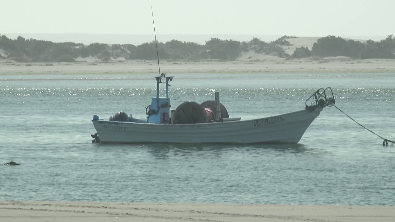 At 0:02, the white fishing boat sits slightly right of center in shallow La Bocana water, with low dunes and scrub forming a muted background and no visible people.
