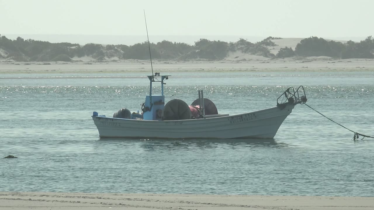 At 0:00, a small white fishing boat floats in shallow water off La Bocana, Baja California Sur, with dunes behind it and no visible people.