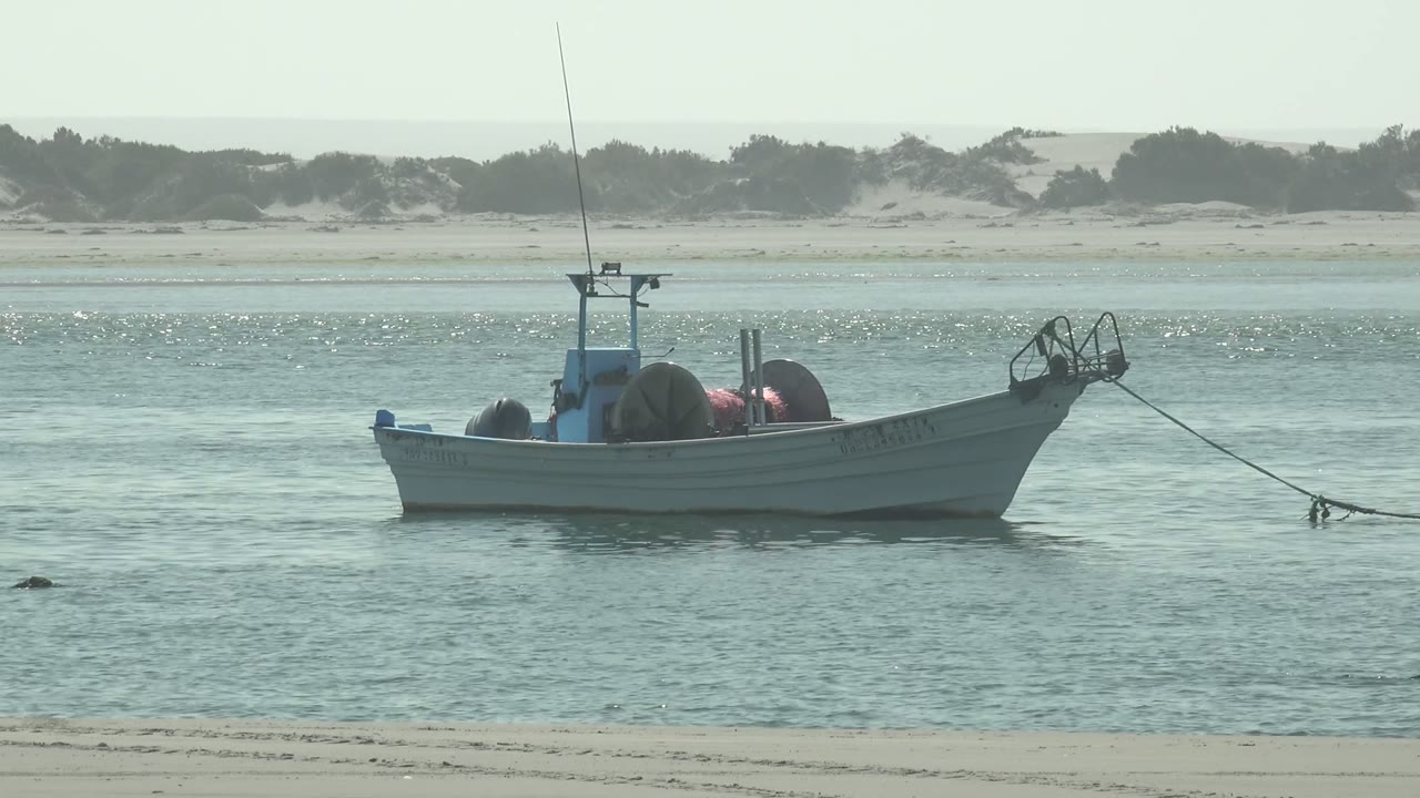 Small white and blue boat anchored in shallow water off La Bocana, Baja California Sur, with beach sand below, dunes behind, and no visible people.