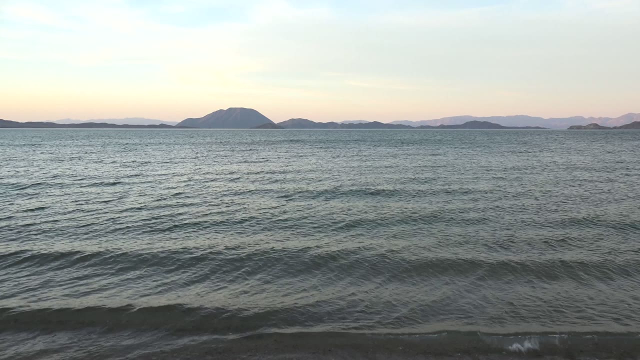 Wide view of Bahia De Los Angeles water with low waves approaching the shore, distant island silhouettes on the horizon, soft pale sky, and no visible people.