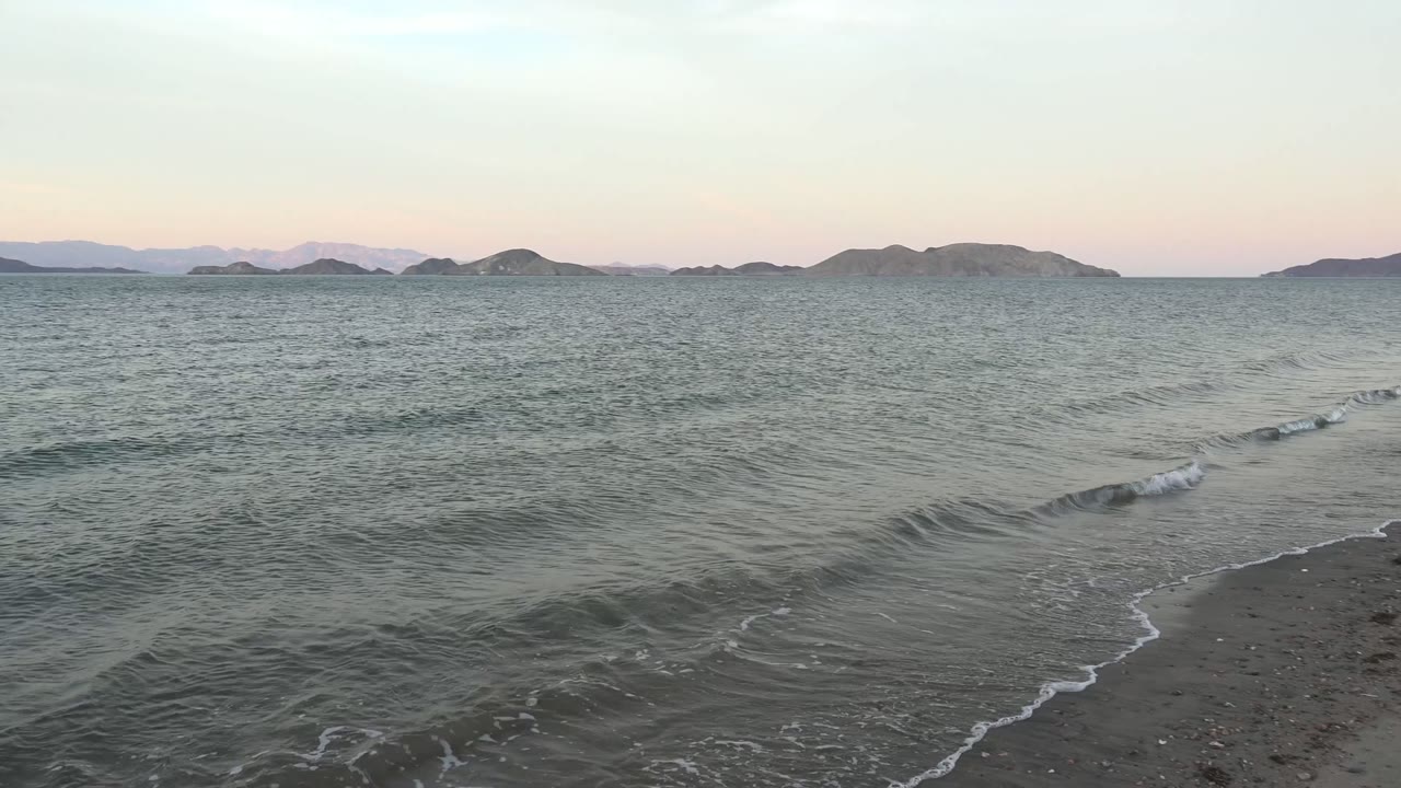 Gentle waves approach the sandy shoreline at Bahia De Los Angeles, Baja California Sur, with distant islands under a pale twilight sky and no visible people.