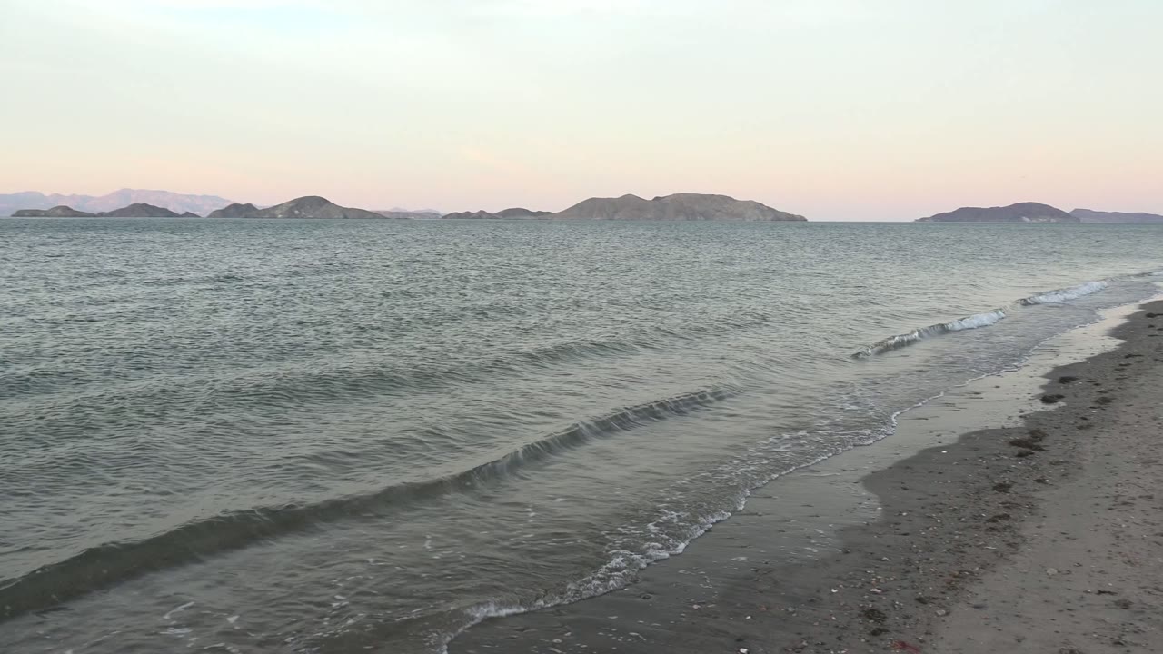 Foamy surf reaches the sandy beach at Bahia De Los Angeles, while open water stretches toward distant island shapes beneath soft evening light and no visible people.