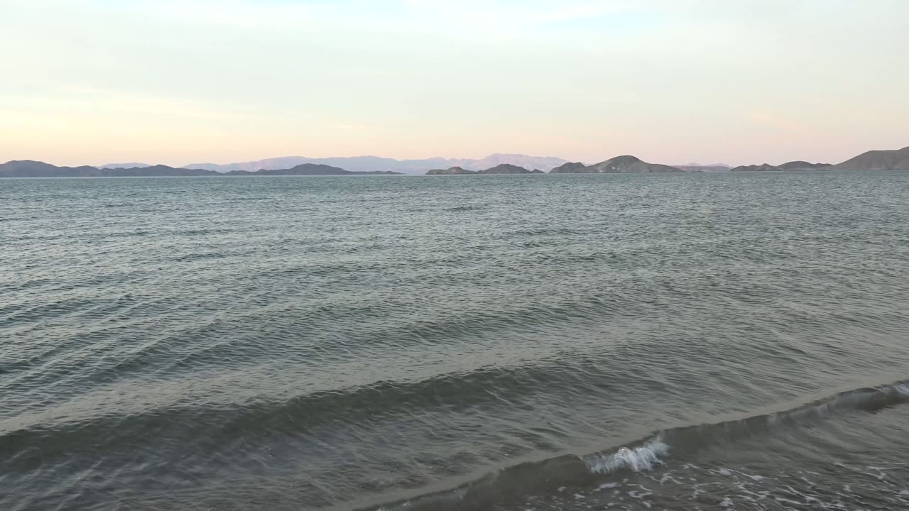 Bahia De Los Angeles shoreline at 0:00 with gray green water, a small wave forming near the beach, pale twilight sky, distant island silhouettes, and no visible people.