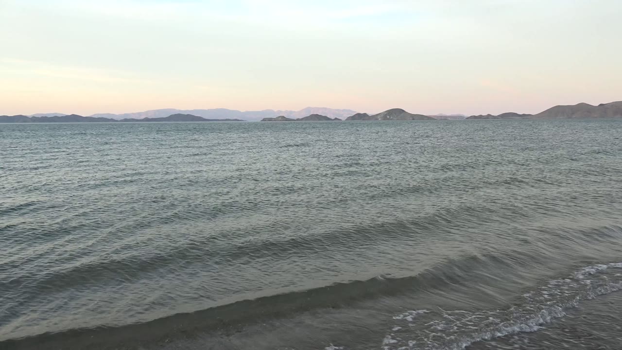 Low surf rolls toward shore beneath a pale sky and island horizon.