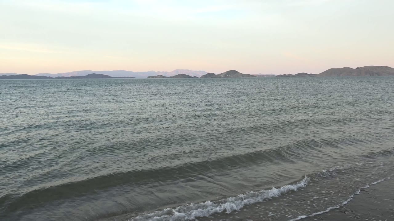 Bahia De Los Angeles at 0:02 with a darker wave ridge crossing the foreground water, fine ripples behind it, muted island shapes along the horizon, and no visible people.