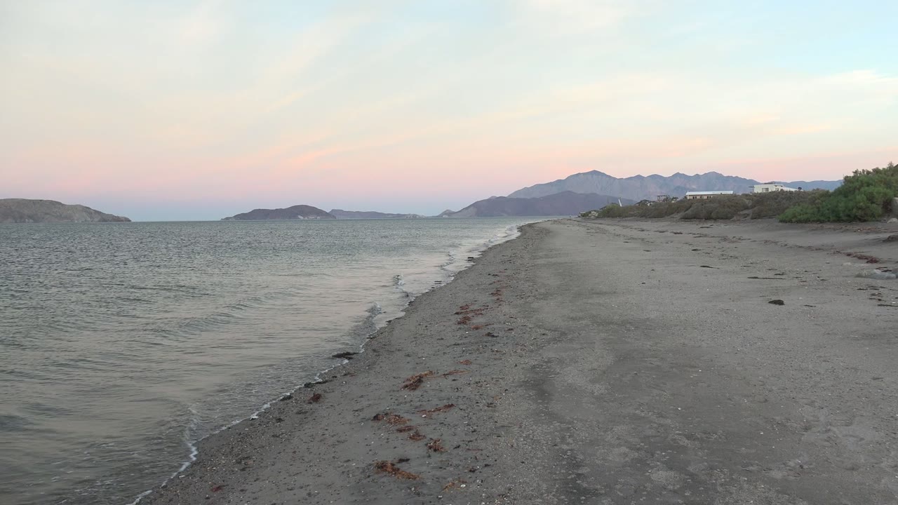 Opening frame of Bahia De Los Angeles, Mexico, showing a sandy shoreline, small waves on the left, distant mountains under a pale pink and blue sky, and no visible people.