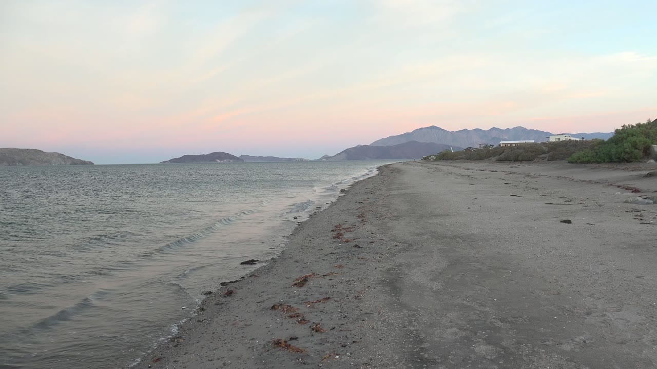 Bahia De Los Angeles shoreline with a thin wave line curling onto the sand, open water to the left, mountains in the distance, low buildings to the right, and no visible people.