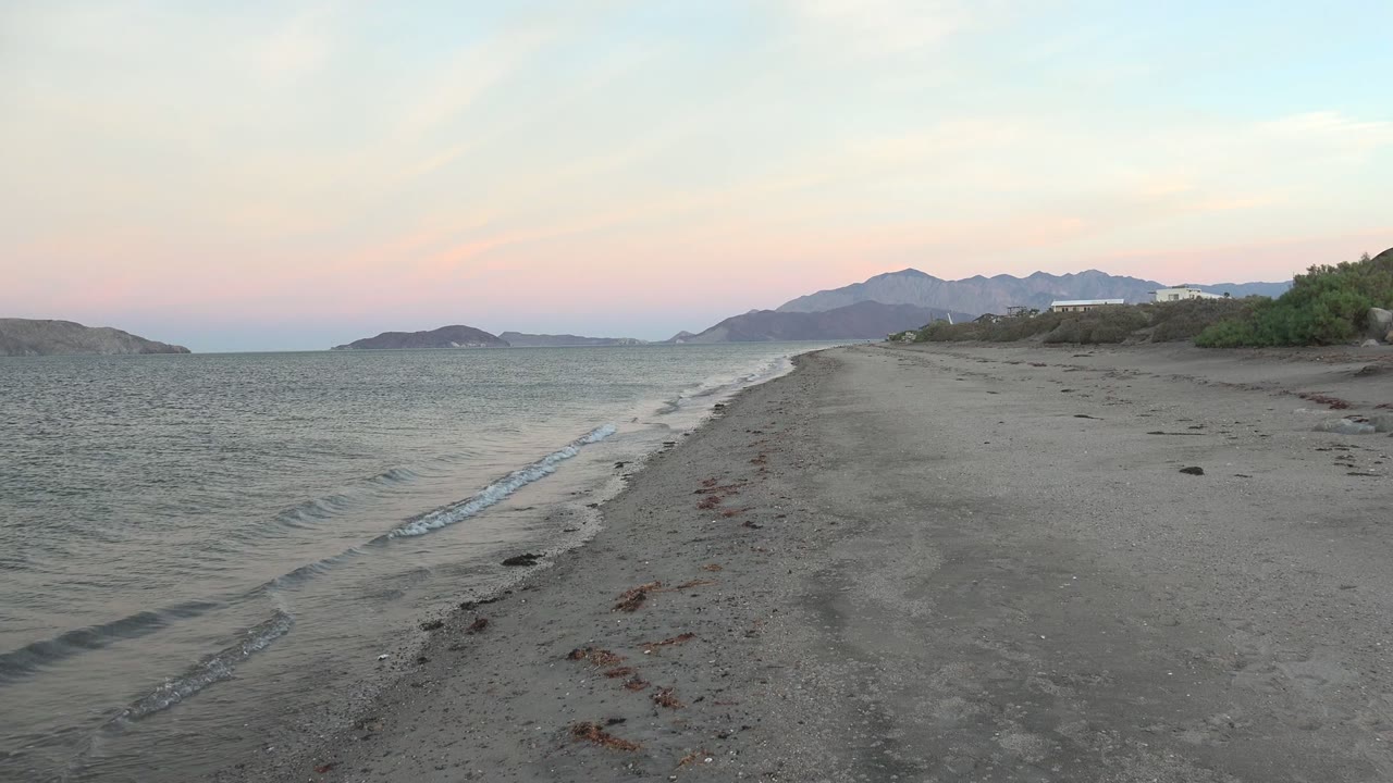 Empty Bahia De Los Angeles beach with shallow waves edging the left side, a broad sandy track through the center, distant headlands and mountains, and no visible people.