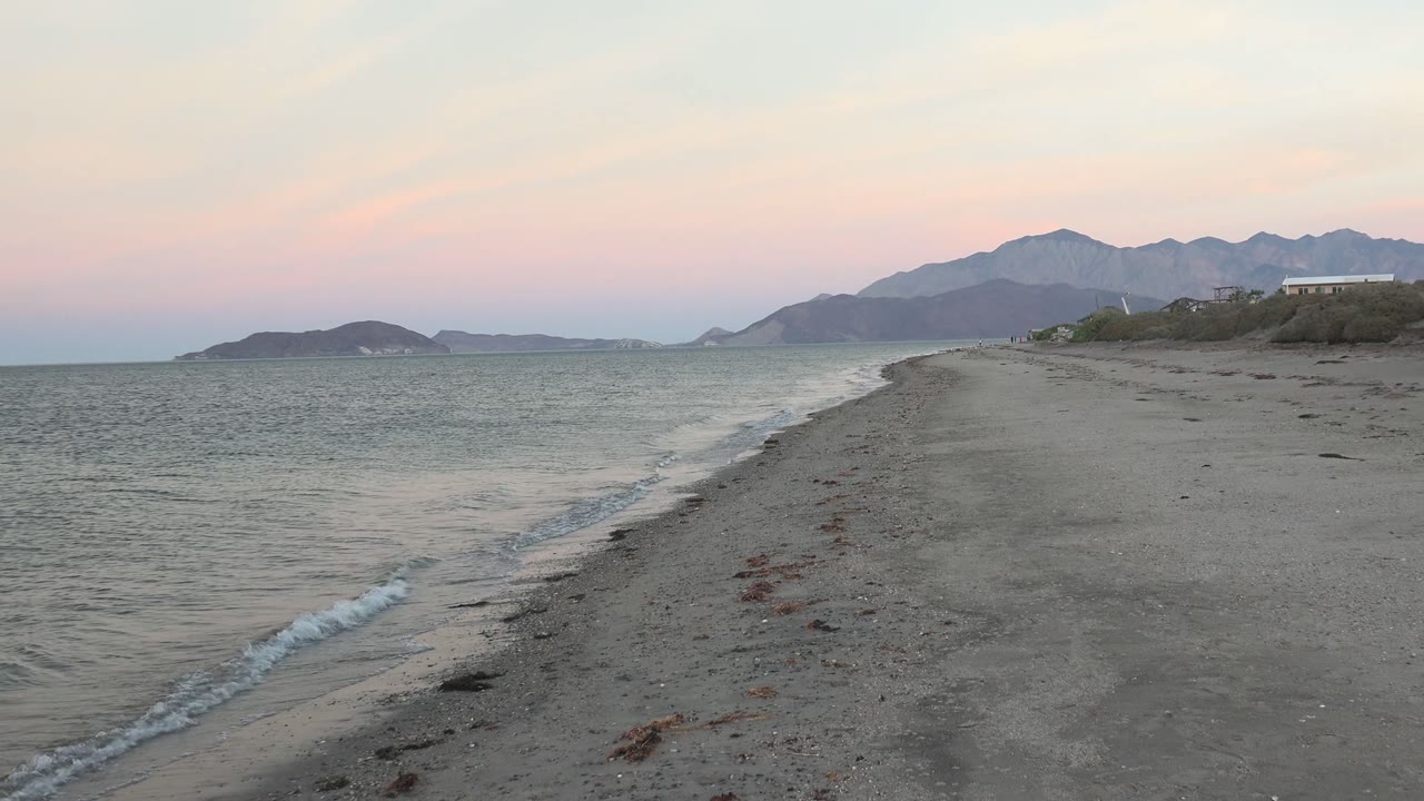 Bahia De Los Angeles shoreline at 0:00 with small waves along the left edge, a sandy spit leading toward distant mountains, and no visible people in this frame.