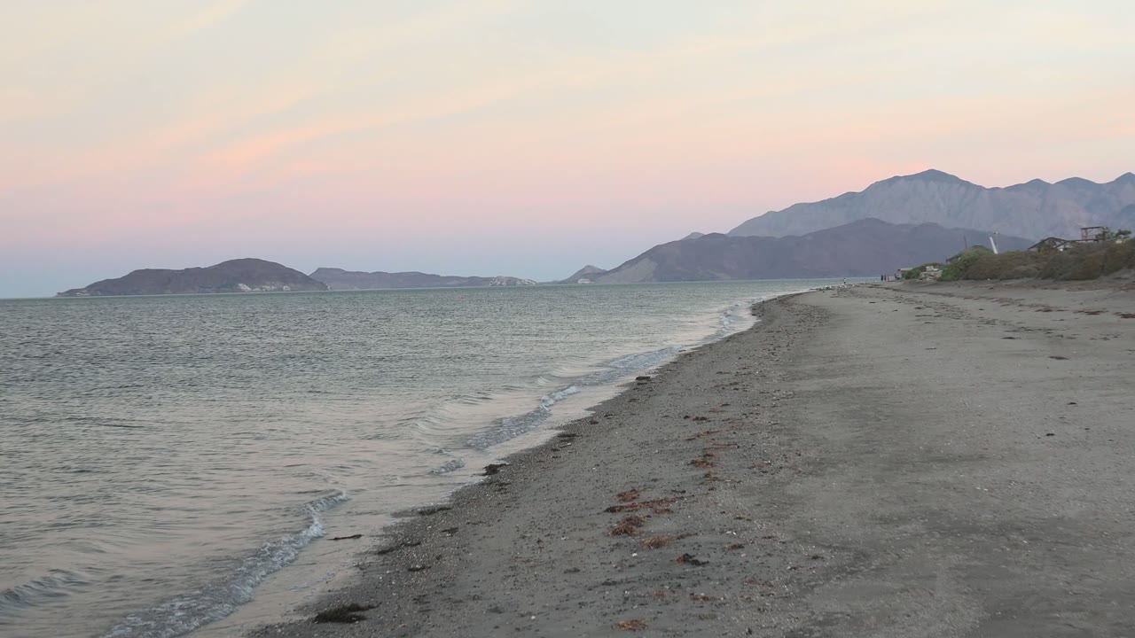 Bahia De Los Angeles shore at 0:02 with a small wave curling near the sand, the beach point running into the frame, and distant mountains beneath pink and blue dusk light.