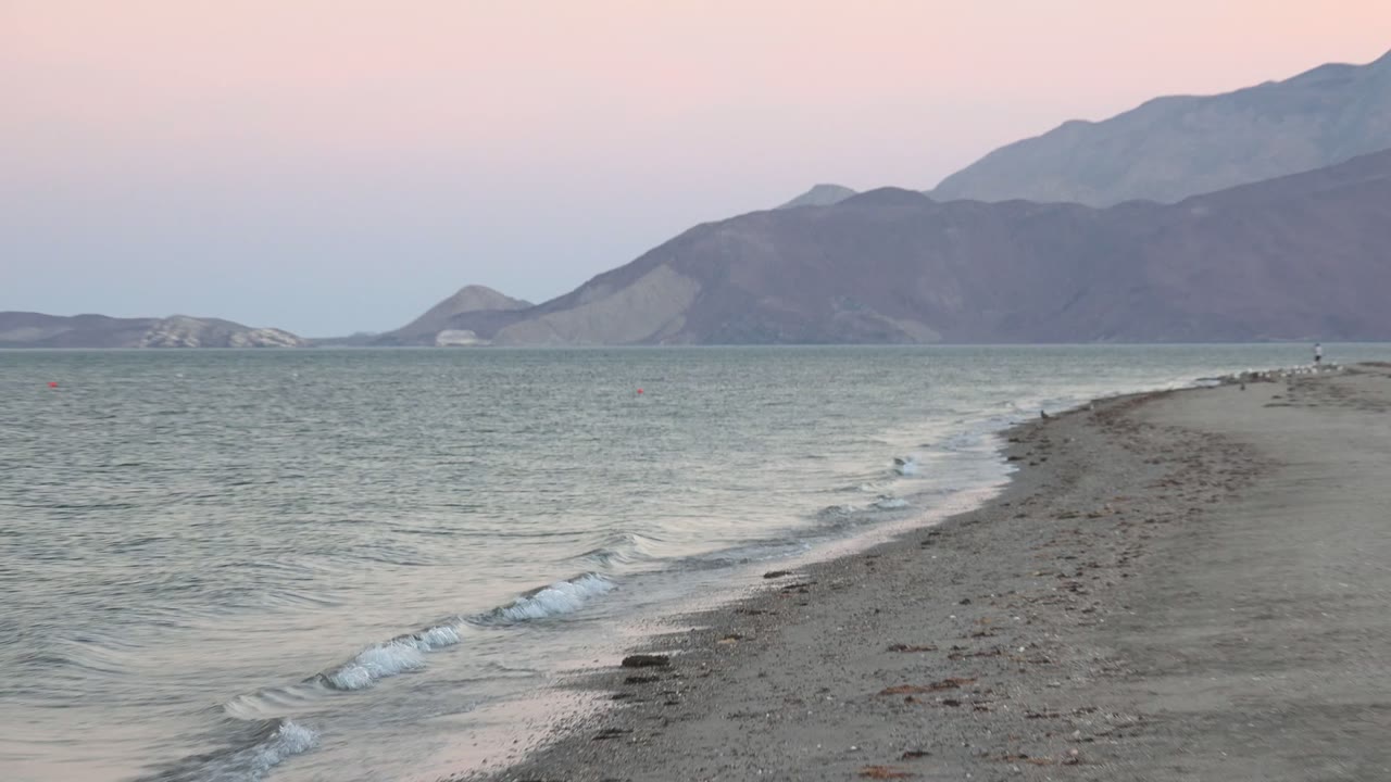 At Bahia De Los Angeles, a rocky sandbar extends into pale water with gray mountains beyond and no visible people.