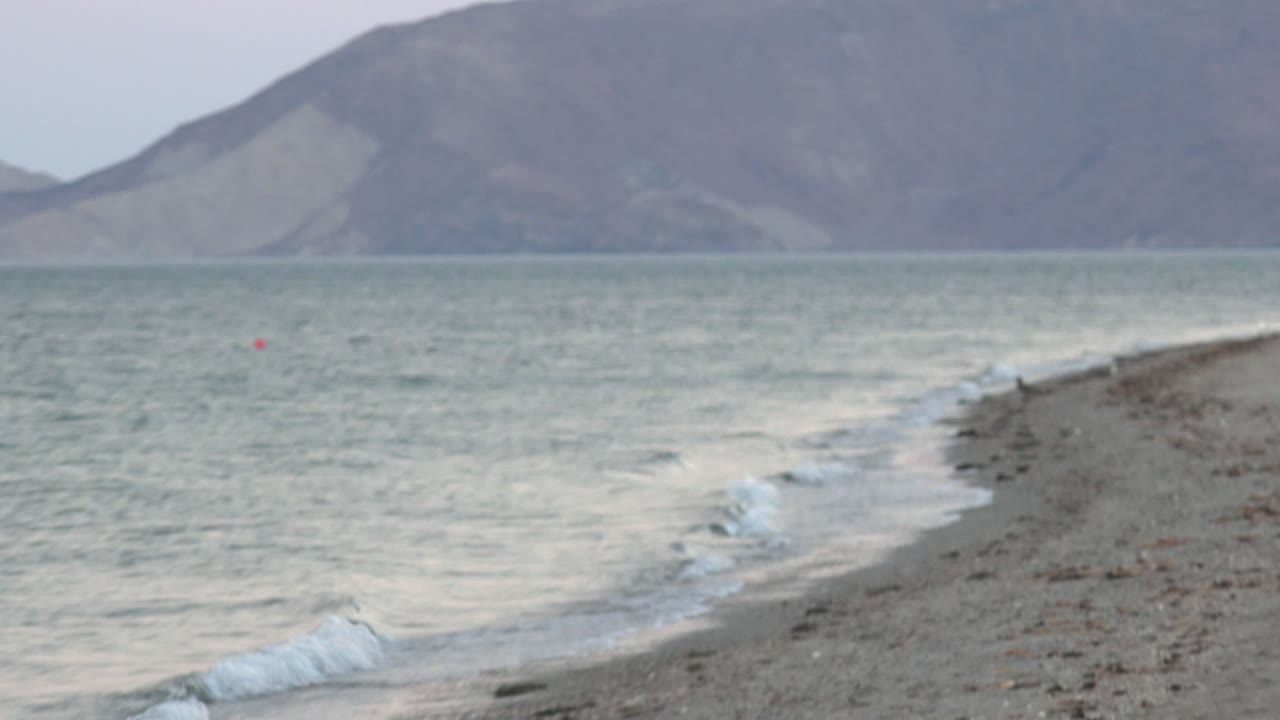 A closer Bahia De Los Angeles view shows low waves curling along the rocky edge, muted green water, gray mountains, and no visible people.
