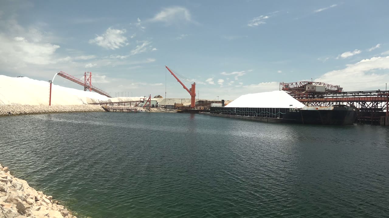 Opening view across rippled water toward an industrial port in Guerrero Negro, Baja California Sur, with white mounds, red cranes, conveyor structures, pale rocks at the lower left, and no visible people.