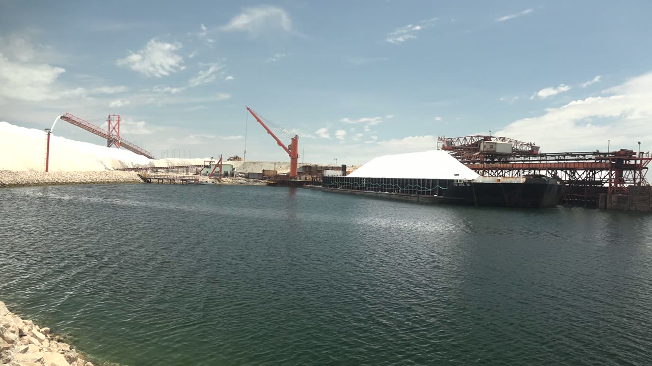 A slightly later view keeps the Guerrero Negro industrial port centered, with green-blue water in front, white mounds to the left and center, red cranes, dark pier framing, and no visible people.