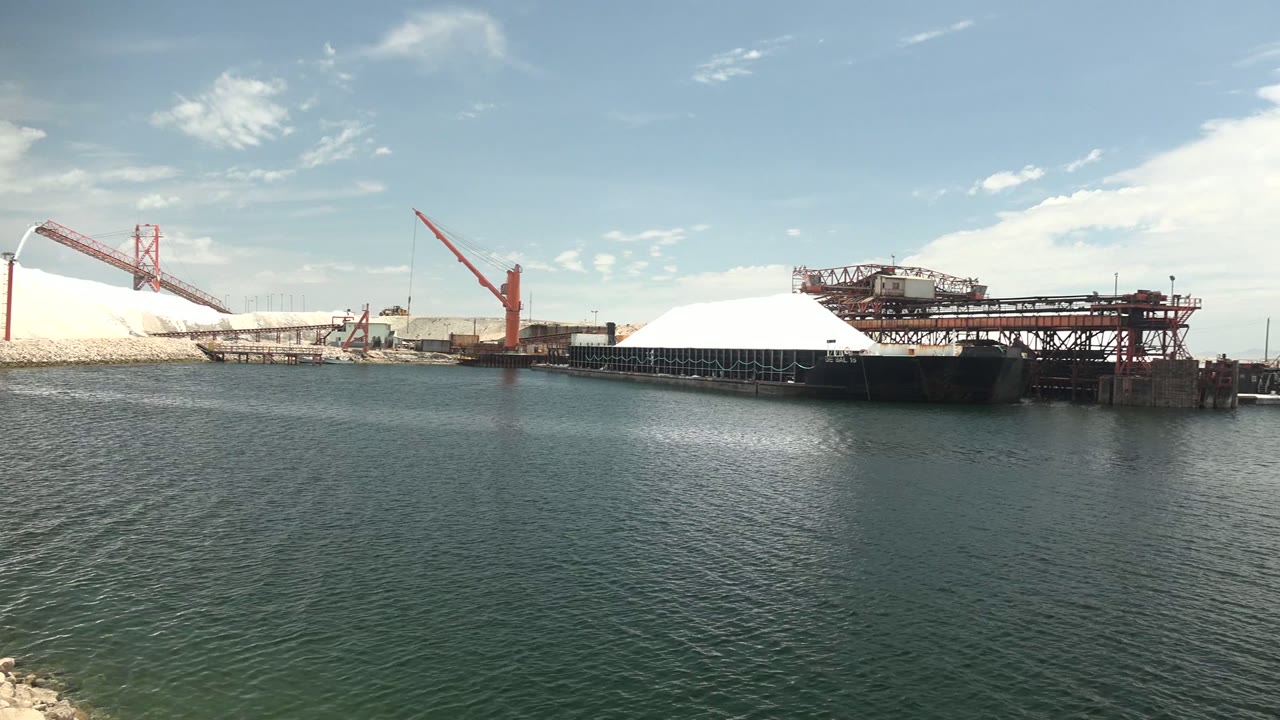 The port remains in a wide low-angle composition in Guerrero Negro, with a broad water foreground, red cranes over white mounds, an elevated conveyor structure at right, and no visible people.
