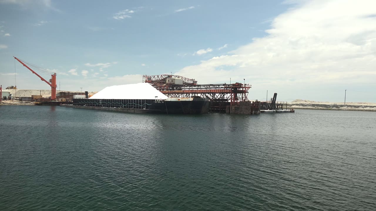 Wide view of an industrial dock in Guerrero Negro with a red crane at left, a white dome structure over the water, calm foreground ripples, and no visible people.