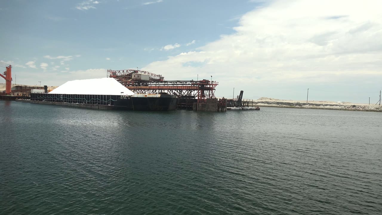 A broad water foreground leads to the white dome and red metal framework on the Guerrero Negro dock, with pale land behind and no visible people.
