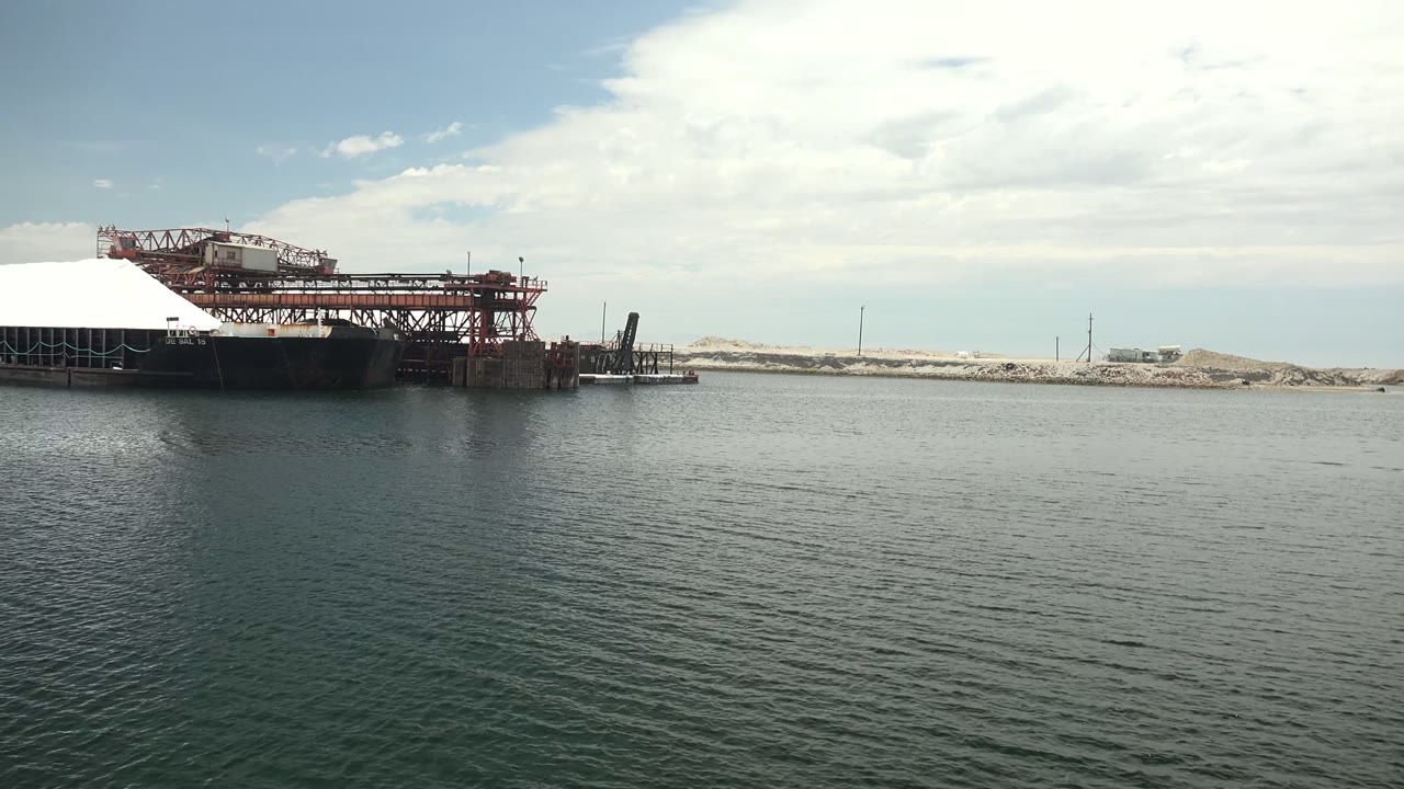 Wide view in Guerrero Negro, Baja California Sur, with rippled water in front, a red industrial dock structure on the left, pale sand piles on the far shore, and no visible people.