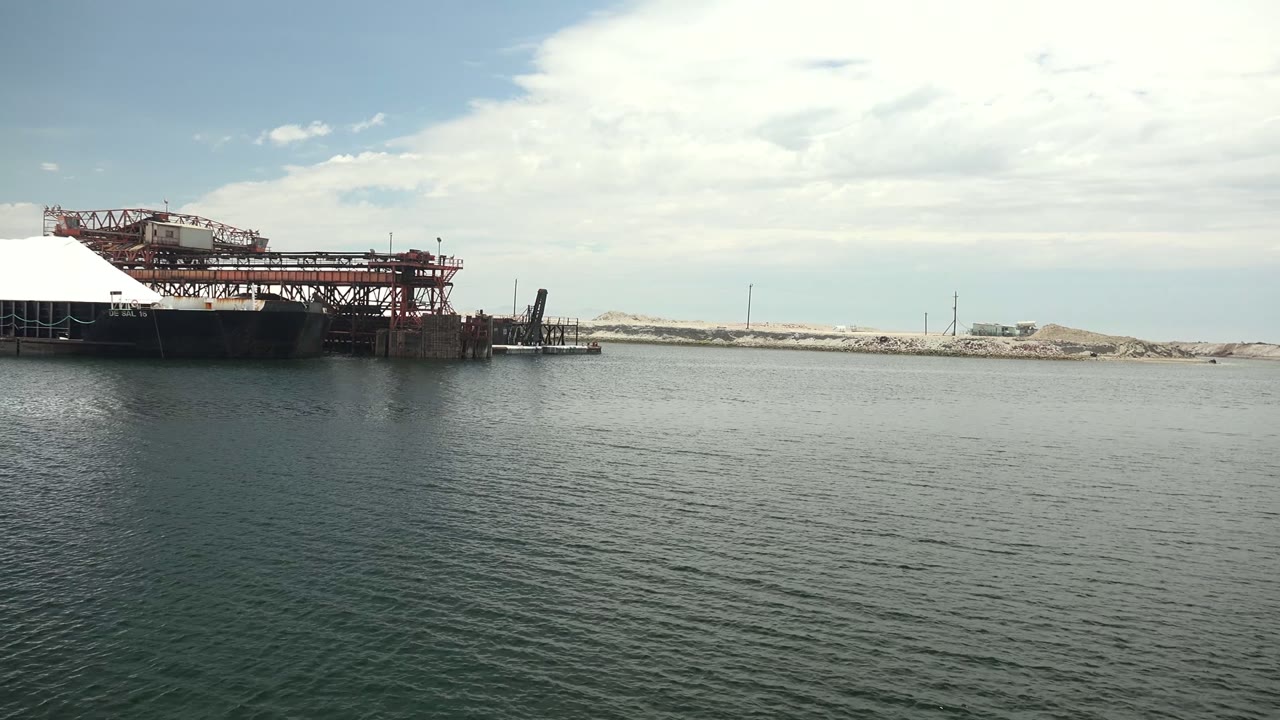 Guerrero Negro waterfront frame with the red metal dock shifted nearer the center-left, dark platform supports over the water, low sand piles behind it, and no visible people.