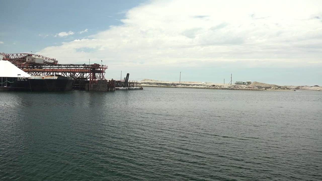 Wide Guerrero Negro harbor view with calm textured water, a red dock or crane structure left of center, pale sand banks across the horizon, scattered poles, and no visible people.