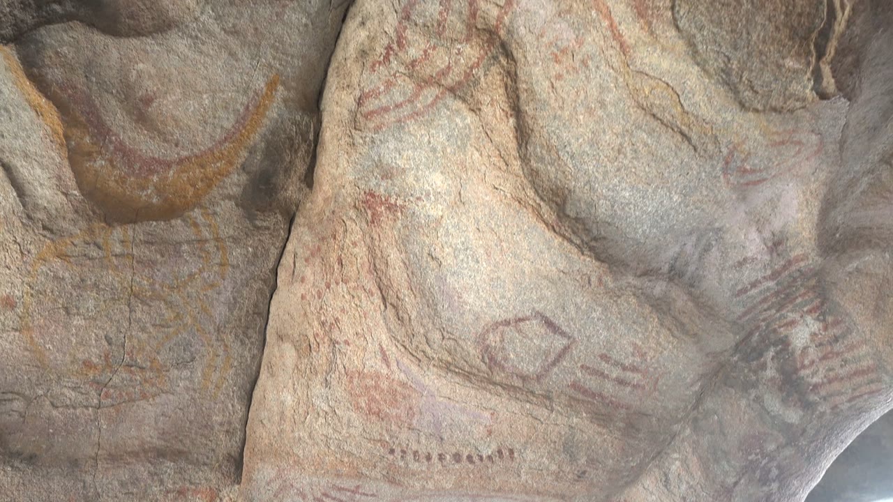 Close view of rock art on a stone face in Santa Rosalia, Baja California Sur, with faded red lines, yellow curved pigment, gray texture, and no visible people.