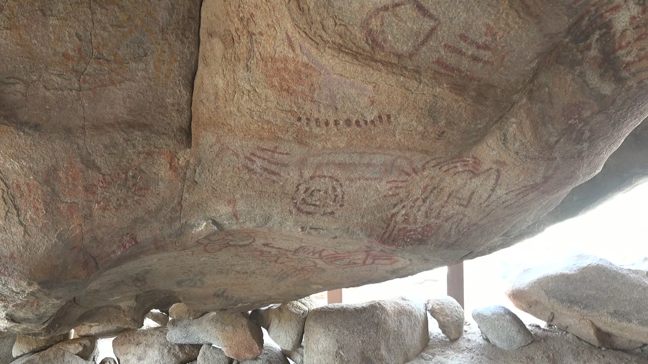 Wide upward view of a Santa Rosalia rock shelter ceiling with faded red markings across rough stone, boulders below the overhang, bright light at the opening, and no visible people.