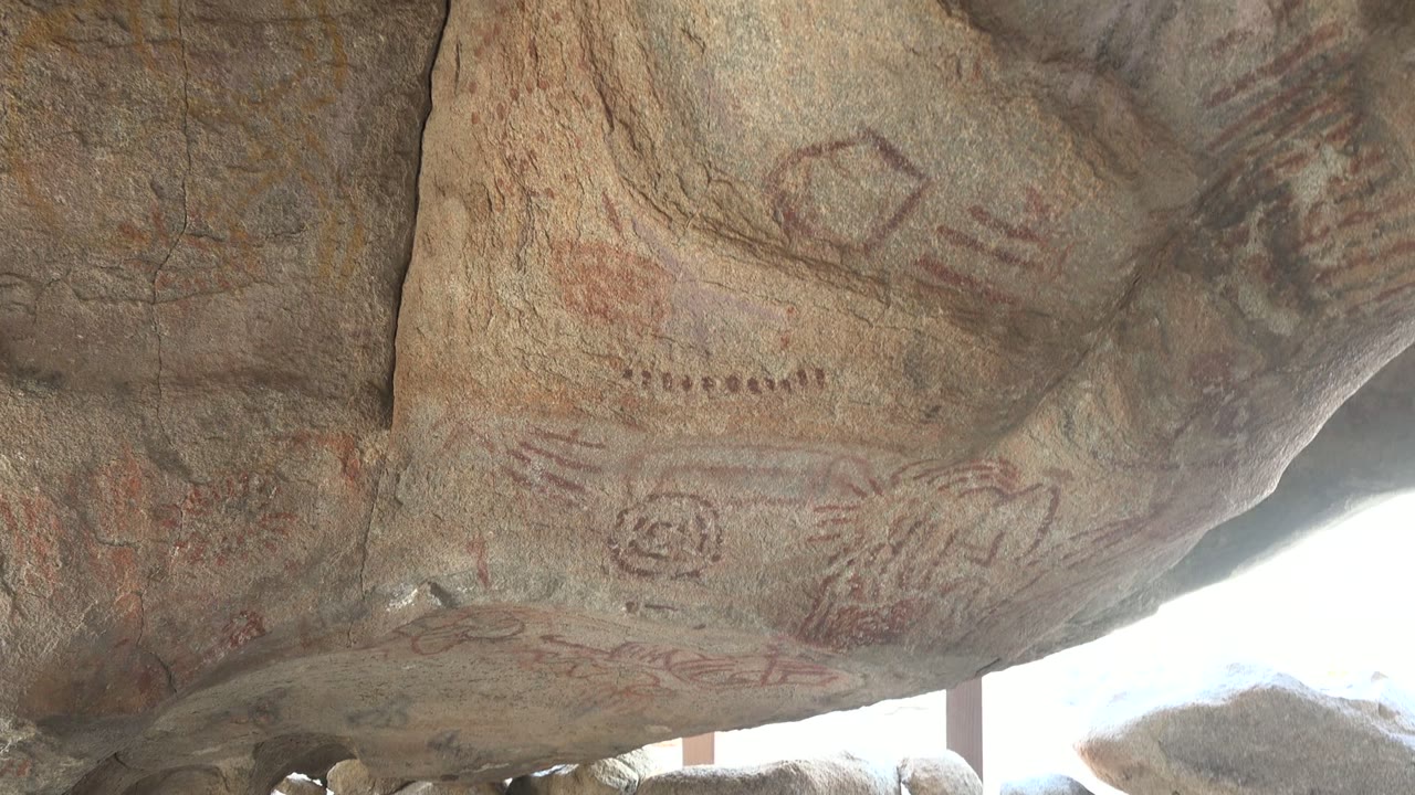 Closer upward composition on the Santa Rosalia overhang, showing faded red and ochre rock art shapes across tan-gray stone, with the bright shelter opening at lower right and no visible people.