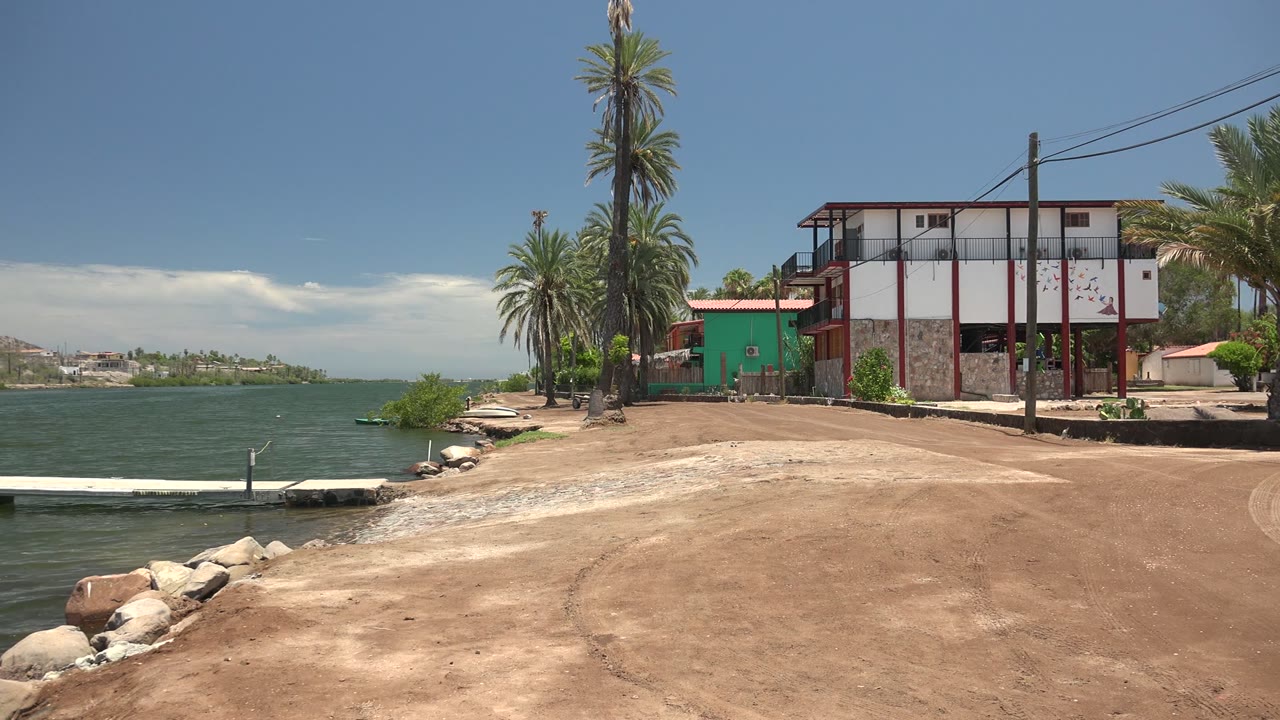 Mulege, Baja California Sur waterfront at the start of the clip, with a small white dock at left, sandy shore in front, palms and a green-and-white building at right, and no visible people.