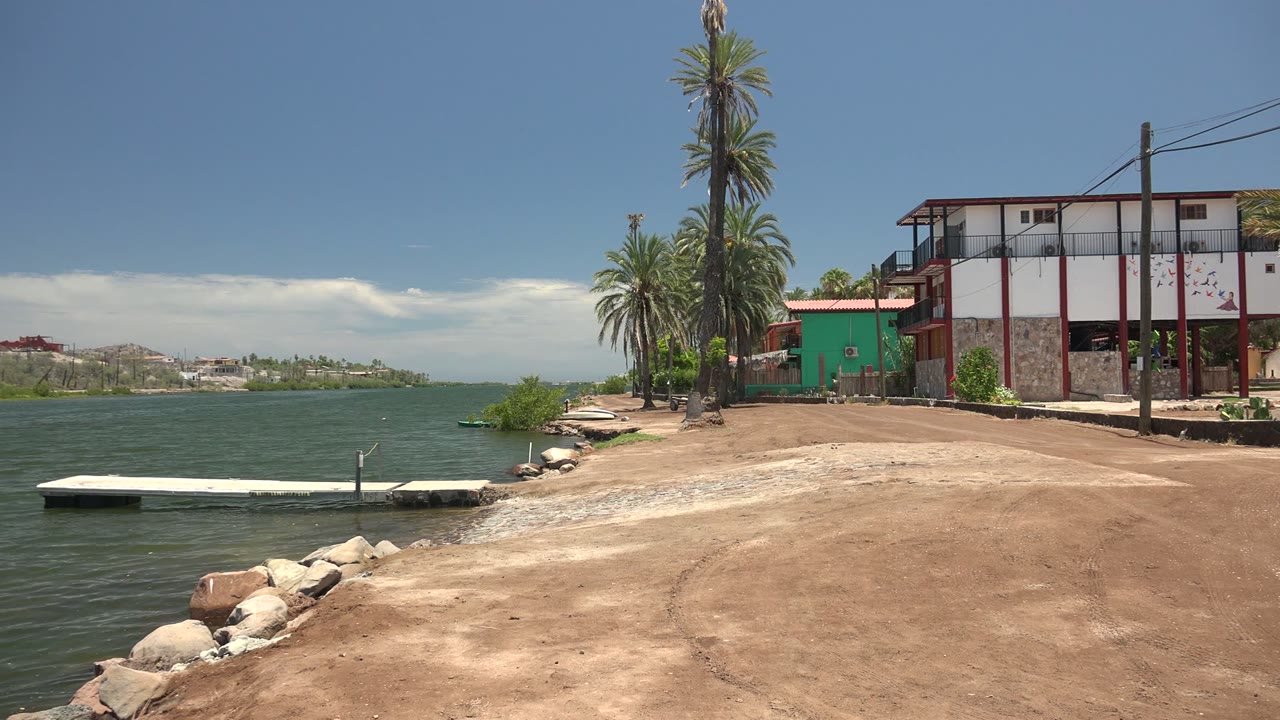 Mulege shore view at 0:02, with ripples around the low dock, foreground rocks along the bank, the sandy access area stretching right, palms, and no visible people.