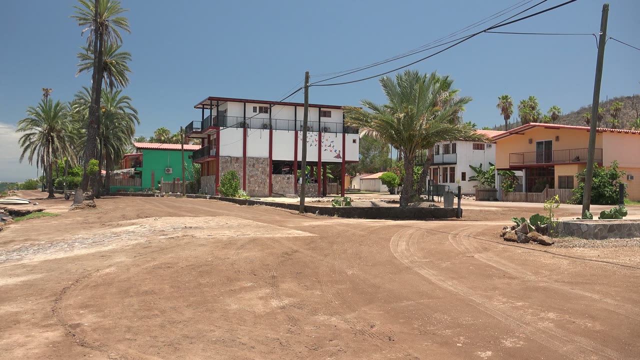 Opening frame of a Mulege, Baja California Sur waterfront road with tall palms, utility lines, a green building, white buildings with red trim, and no visible people.