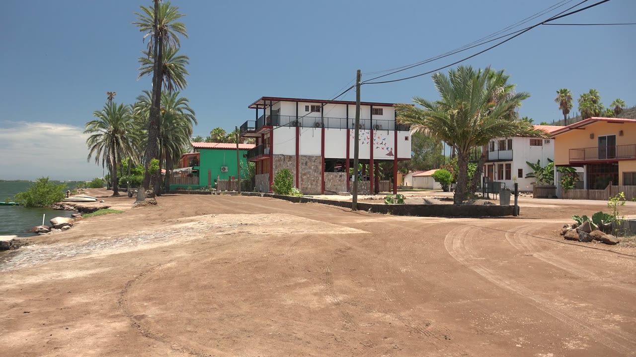 A leftward pan position in Mulege shows the dirt road curving near waterfront homes, palms, low rocks by the water, and no visible people.
