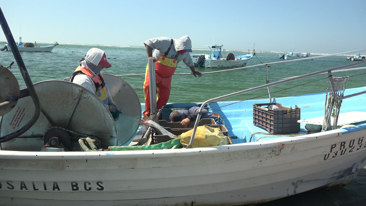 Two fishermen work on a small boat in La Bocana, Baja California Sur, with a net reel in the foreground and other boats on calm water behind them.