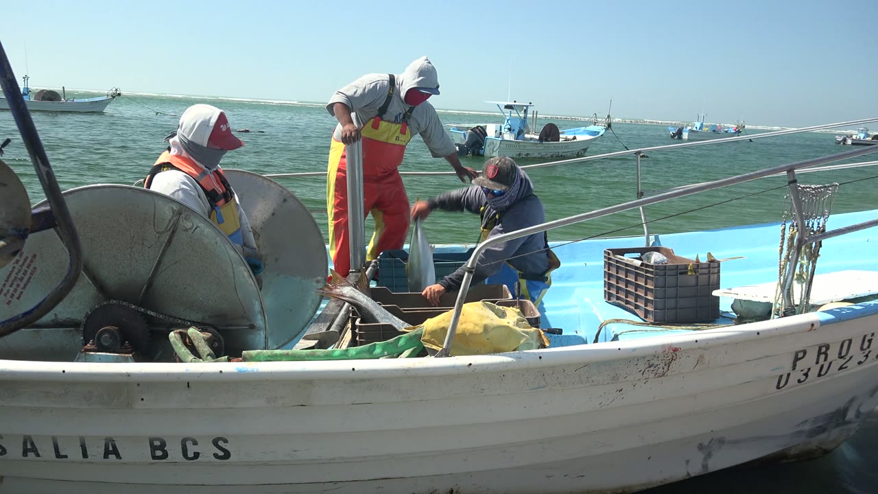 One fisherman stands while another crouches near deck gear on a small boat.
