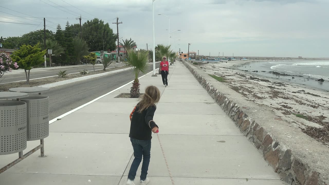 At 0:00 in Punta Abreojos BCS Mexico, a girl is seen from behind on a concrete sidewalk holding a leash, with a man in a red shirt ahead, the ocean to the right, and palms and road to the left.