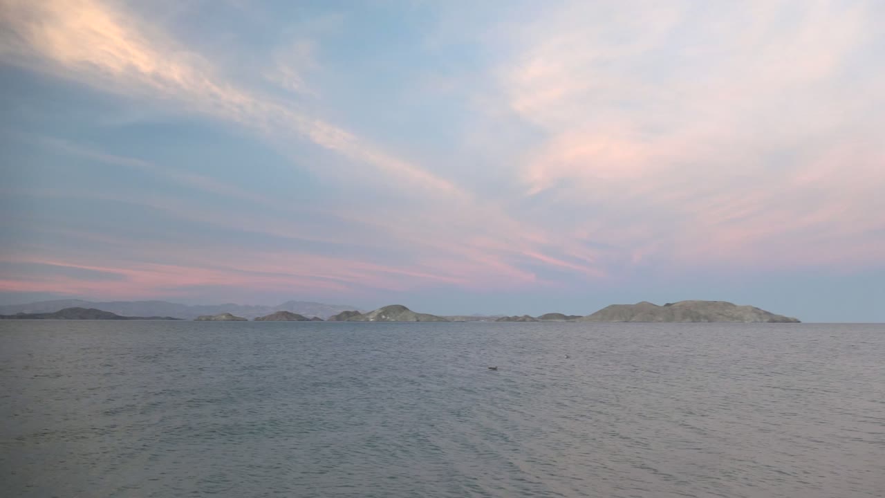 Calm sea in Bahia De Los Angeles, Mexico, with low distant islands on the horizon, pink and blue sunset clouds overhead, and no visible people.