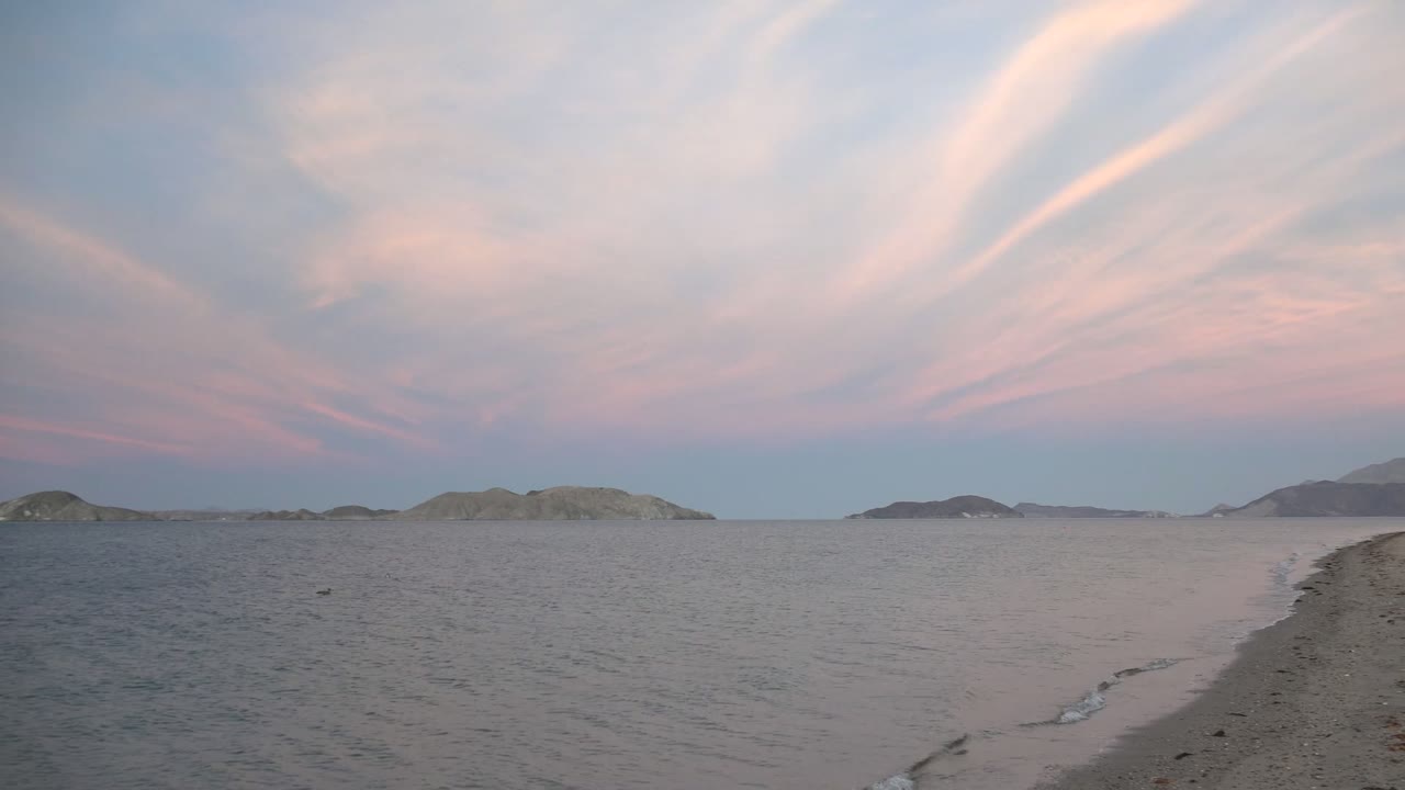 Bahia De Los Angeles beach at twilight with a thin wave line along the sand, distant islands on the horizon, pink-blue cloud bands overhead, and no visible people.