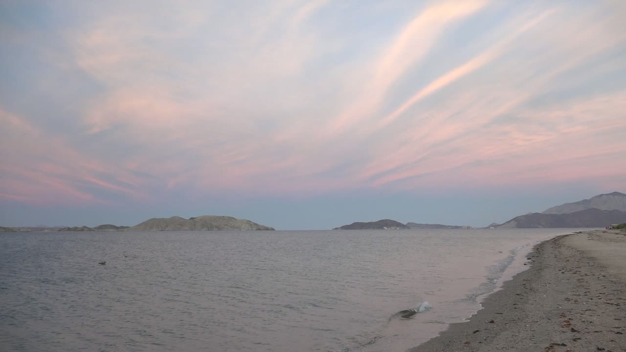 Bahia De Los Angeles beach view at 0:02 with a small wave touching the right edge of the sand, open water to the left, distant landforms on the horizon, and no visible people.