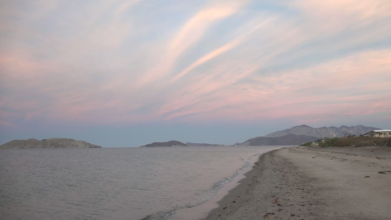 Bahia De Los Angeles sandy beach with calm water on the left, distant island shapes on the horizon, pastel pink clouds above, and no visible people.
