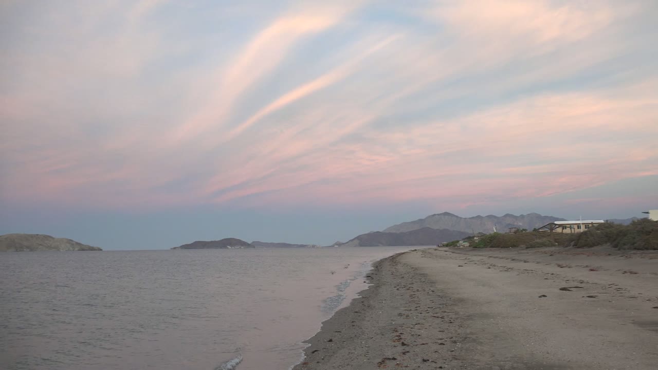 Bahia De Los Angeles beach view with more open sand at right, calm water at left, distant mountains on the horizon, streaked pink clouds, and no visible people.