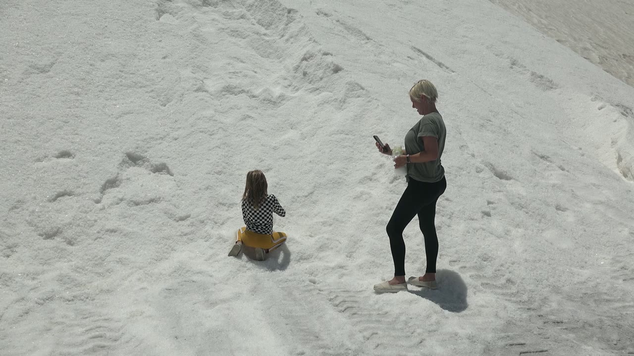 At 0:00 in Guerrero Negro, Mexico, a child sits on a yellow sled on the white salt flat while an adult in a gray shirt stands to the right holding a phone.