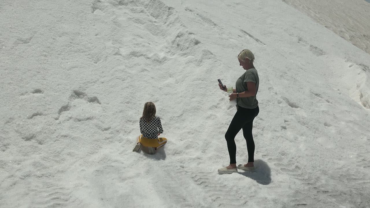 Child remains seated on the sled as an adult stands nearby on white salt.
