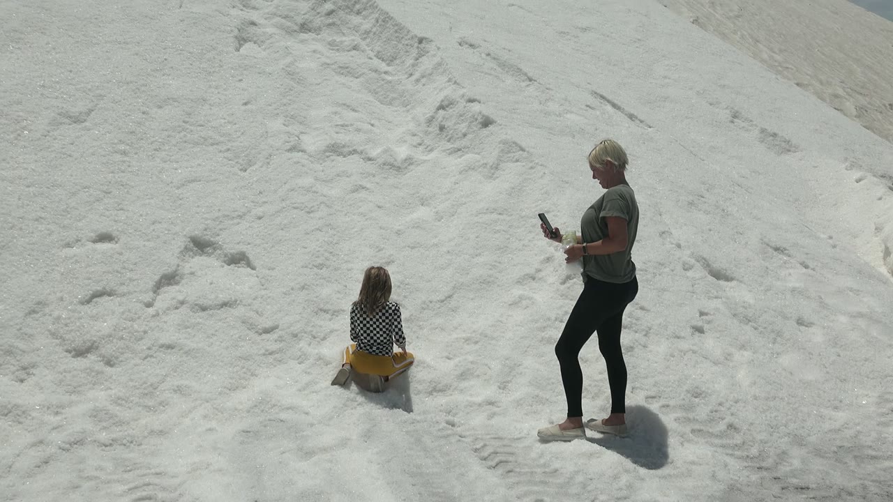 At 0:02 in Guerrero Negro, Mexico, the child remains low on the yellow sled while the adult stands with a phone, both surrounded by pale salt mounds and harsh sunlight.