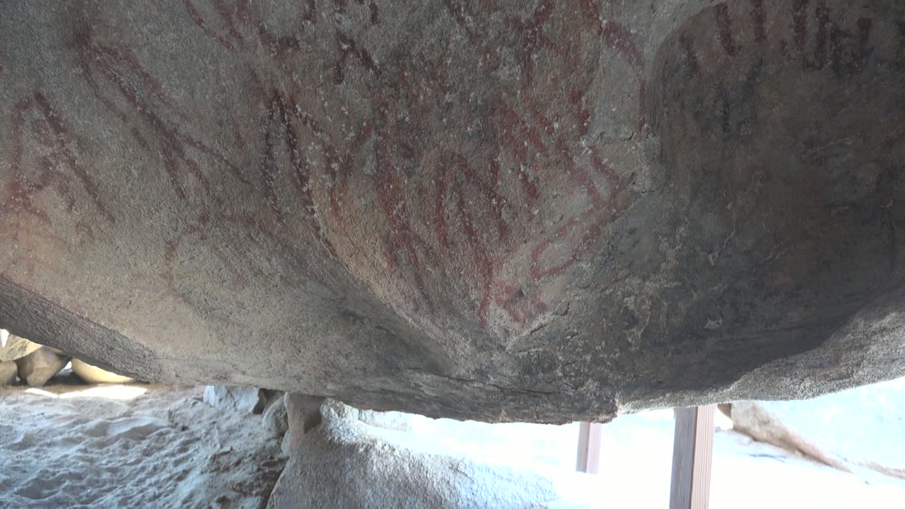 Early frame of red pictographs and faint symbols across a gray rock shelter ceiling in Santa Rosalia, Baja California Sur, with bright light below and no visible people.