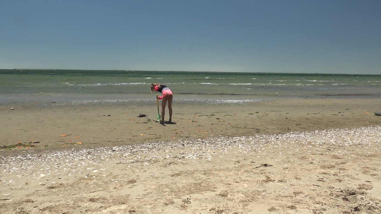 Person in pink remains bent over on the beach with waves behind.
