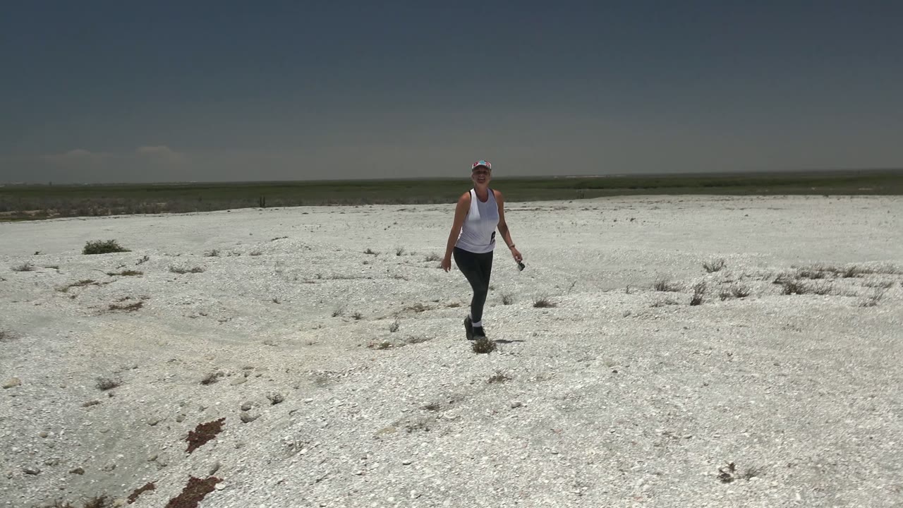 A woman begins walking toward the camera across a white rocky desert landscape in San Carlos, Mexico, with sparse brush and a flat horizon behind her.
