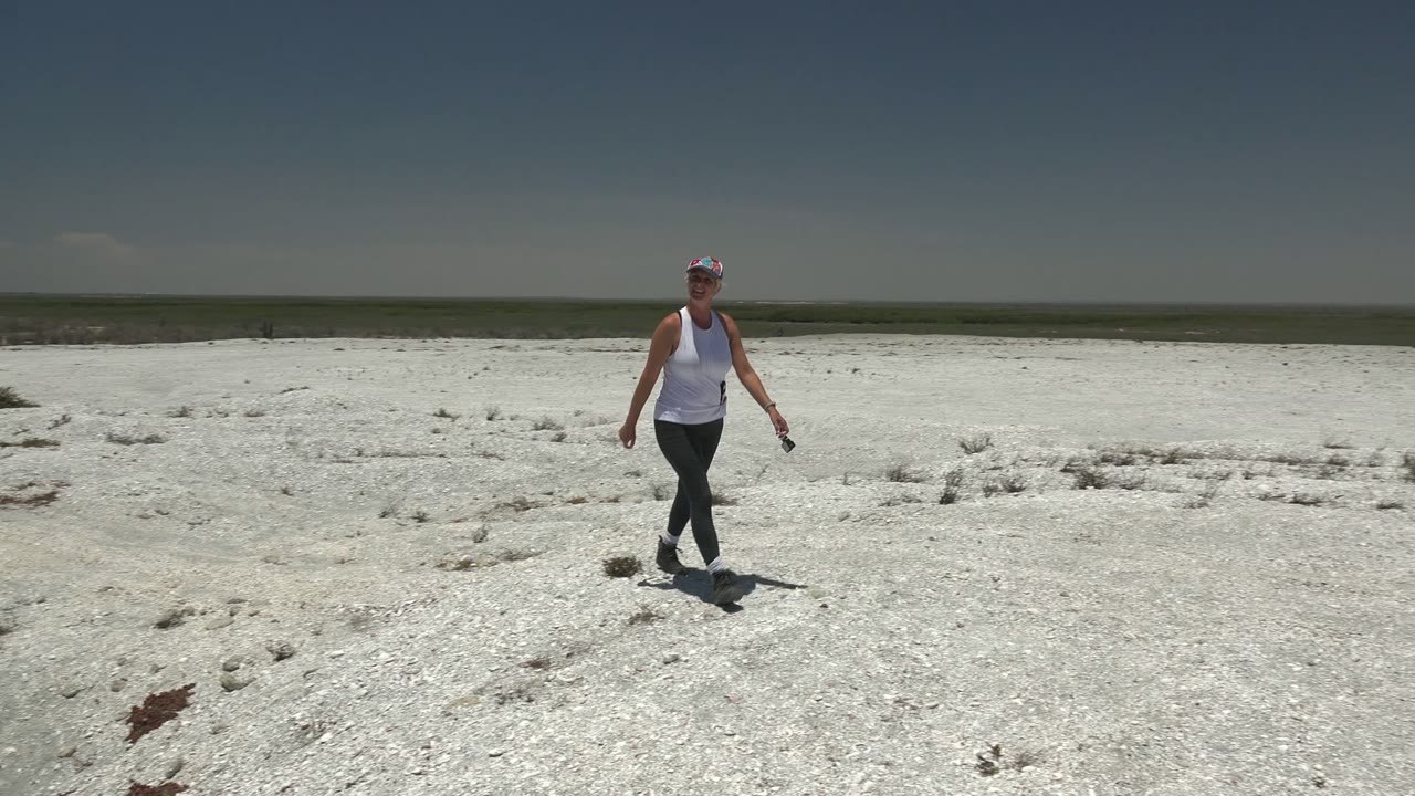 The woman moves closer across the pale rocky ground in San Carlos, Mexico, with low scrub scattered across the bright desert surface.