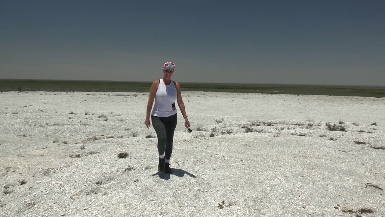 A woman in athletic clothing walks toward camera across the flat white desert in San Carlos, Mexico, holding a small dark object at her side.